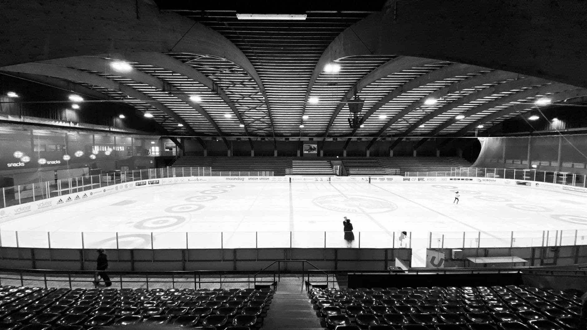 Black and white view of an empty indoor ice rink with a few spectators observing.